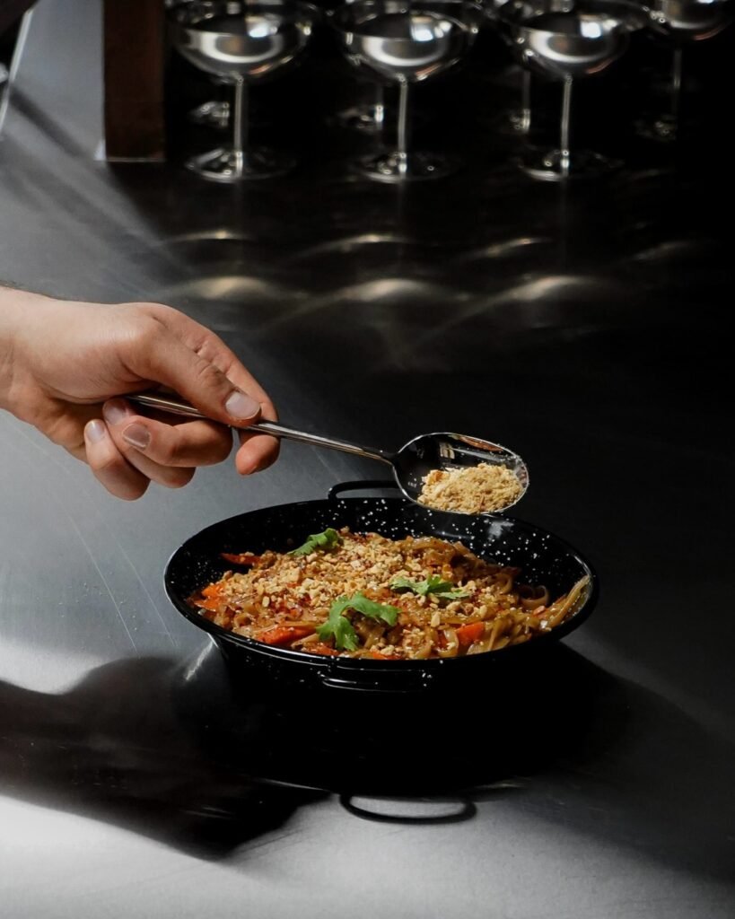 ذا تاي تيبل The Thai Table: طبق نودلز تايلندي يُحضّر على الطريقة التقليدية، ويُزيَّن بالفول السوداني المجروش والكزبرة الطازجة، ليجمع بين النكهات الغنية والتقديم الأنيق