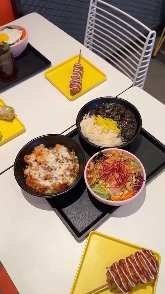 أومو أومو : مجموعة أطباق كورية : نودلز بصوص الفاصوليا السوداء، طبق مخبوز بالجبن، سلطة كورية ملوّنة، أرز مع بيض، وكورندوغ مغطى بالكاتشب والمايونيز. مائدة مليئة بألوان ونكهات آسيوية جذابة.